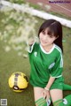 A woman in a green soccer uniform sitting next to a yellow soccer ball.